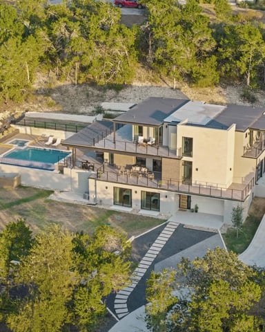 Aerial view of modern multi-level home with pool and hot tub surrounded by dense forest and driveway