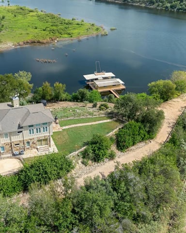 Waterfront house with stone and wood exterior on river bluff, moored boat, manicured lawn, and forested shoreline.