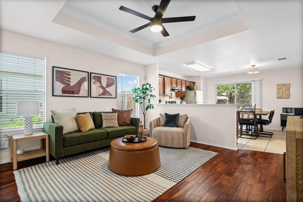 Open-plan living room with green sofa, beige armchair, round brown ottoman, and wood flooring, opening to kitchen and dining area