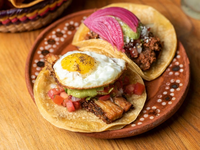 Two breakfast tacos on a terracotta plate with fried egg, pico de gallo and guacamole